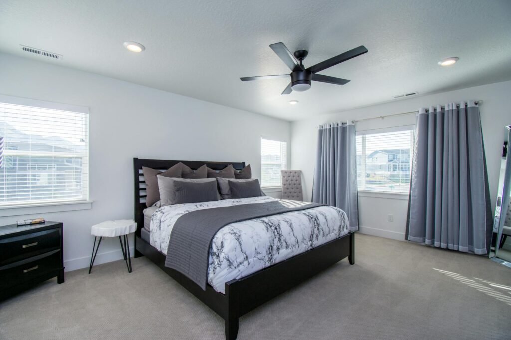 A tidy neutral bedroom with blackout curtains, a fan, and minimal electronics